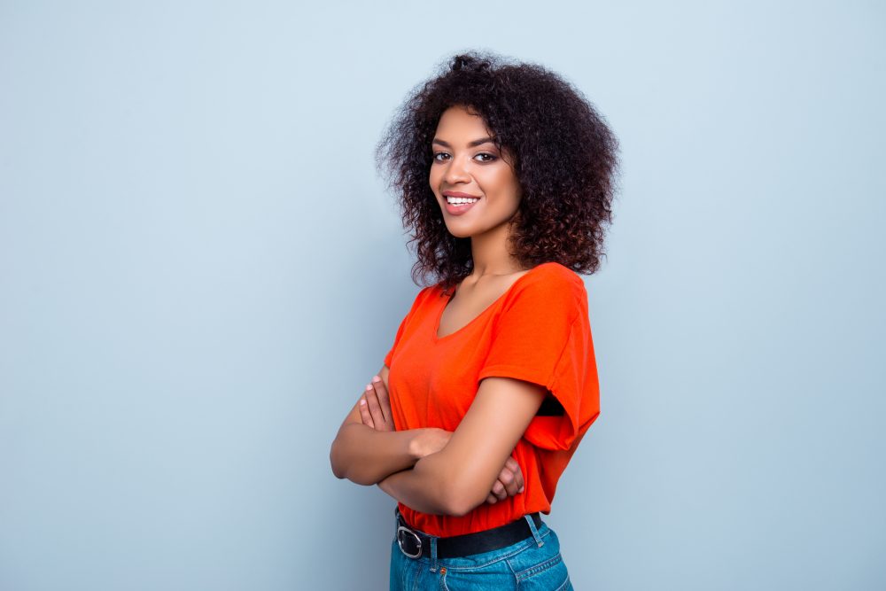 Portrait of joyful cute woman with plump lips modern hairdo in orange t-shirt holding hands crossed looking at camera isolated on grey background