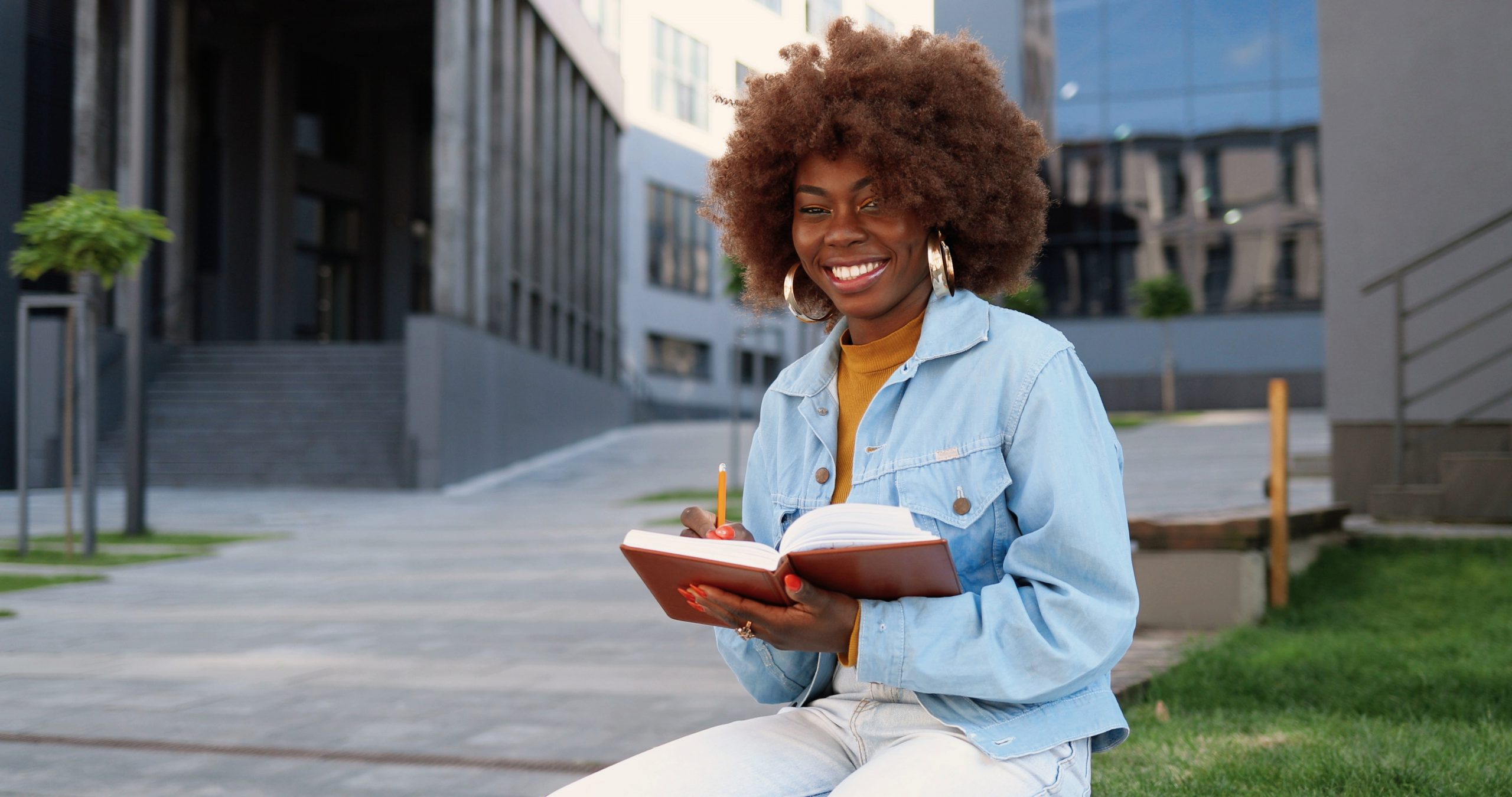 Portrait of young curly and pretty African American woman sitting on bench, reading textbook, making marks and smiling to camera.